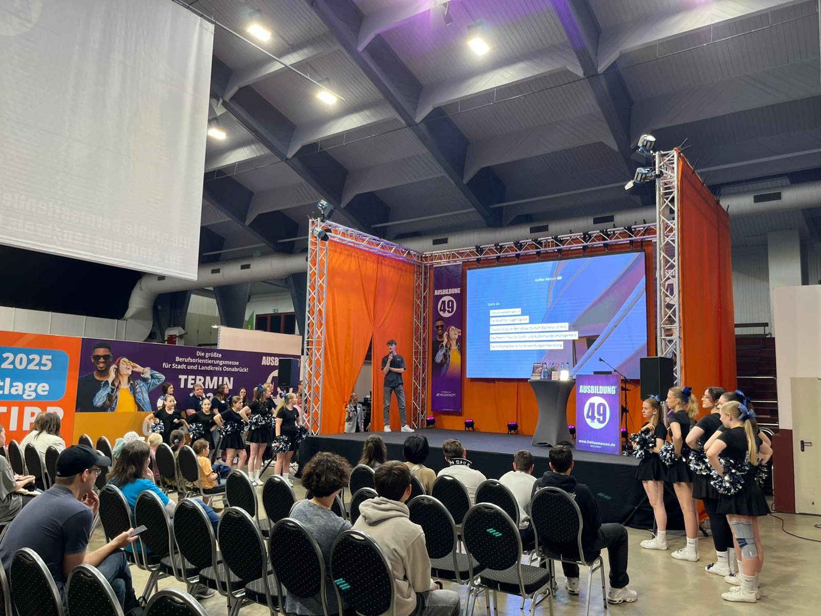 AUSBILDUNG event stage with speaker, cheerleaders, audience. Screen displays "Ausbildung 49" logo. d63a3d2a-6ac2-4787-a991-7c819e705930.jpg
