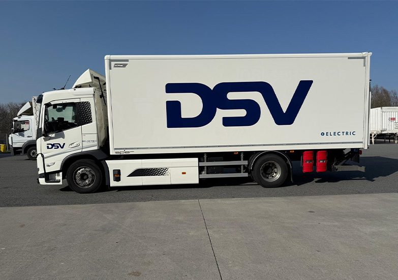 Schenker E-Truck: White electric truck with a blue DSV logo on the side. The truck is parked on a concrete surface under a blue sky.