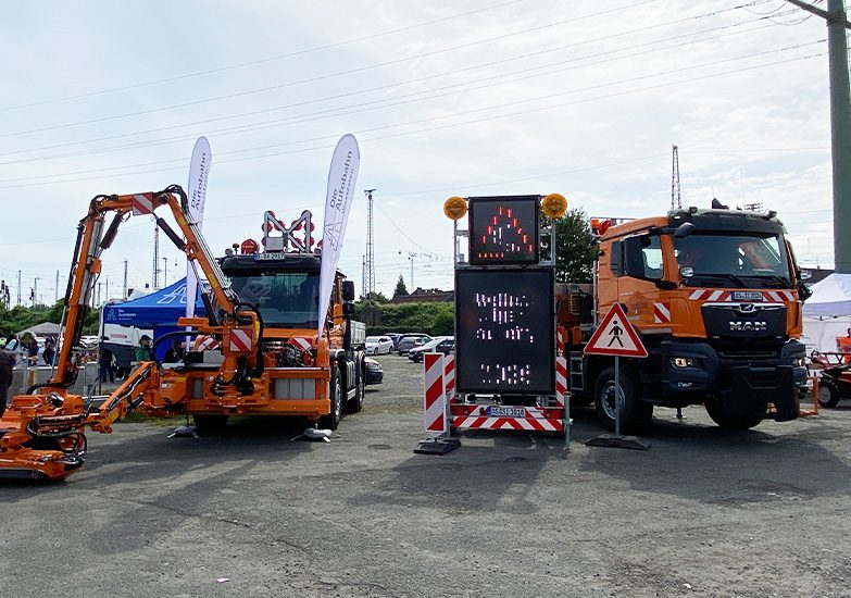 Die Autobahn Unimog: Zwei orangefarbene Nutzfahrzeuge, eines mit Mäharm, das andere mit LED-Verkehrswarntafel, auf einem Parkplatz.